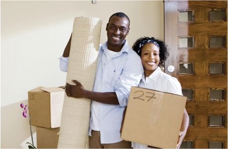 A smiling couple stand by a wooden door, holding moving boxes.