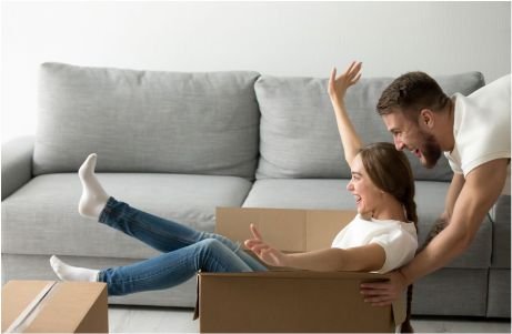 Un hombre empuja juguetonamente a una mujer sonriente sentada en una caja de cartón.